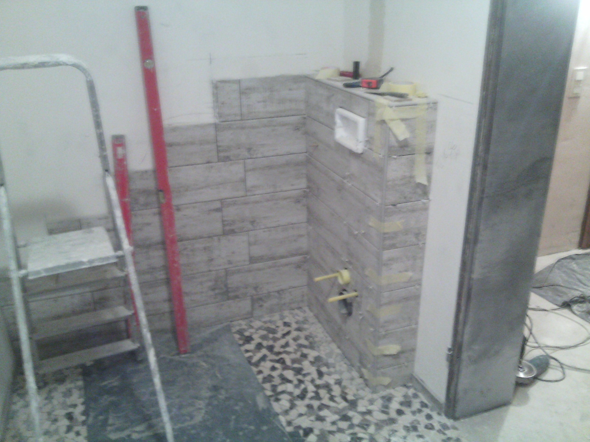 Łazienka w trakcie układania płytek imitujących drewno na ścianie i mozaiki na podłodze, widoczna drabina, poziomica i narzędzia.