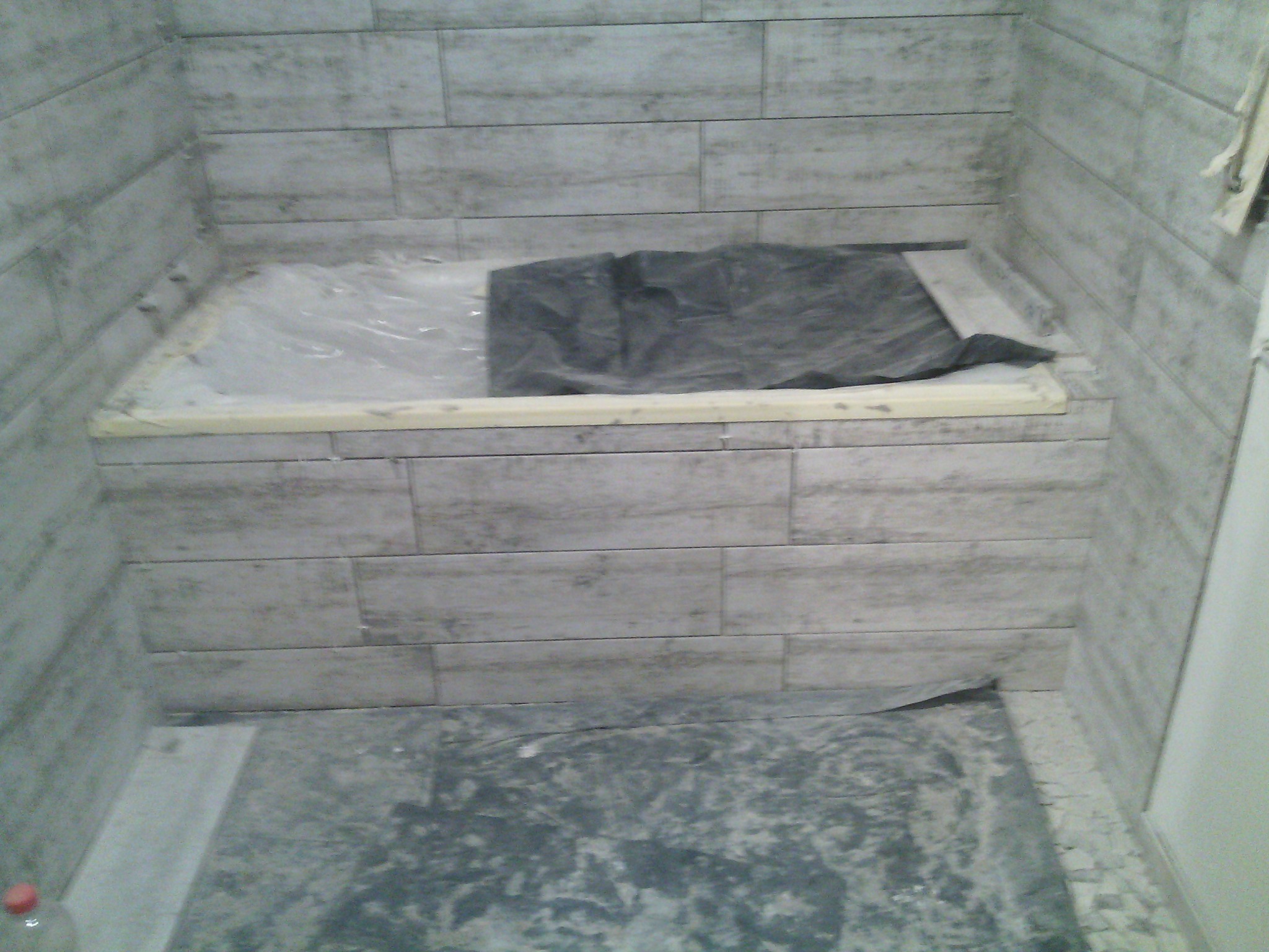 Przygotowanie do wykończenia obudowy wanny płytkami imitującymi drewno, zabezpieczona folią i taśmą malarską, podłoga częściowo zakryta folią ochronną.