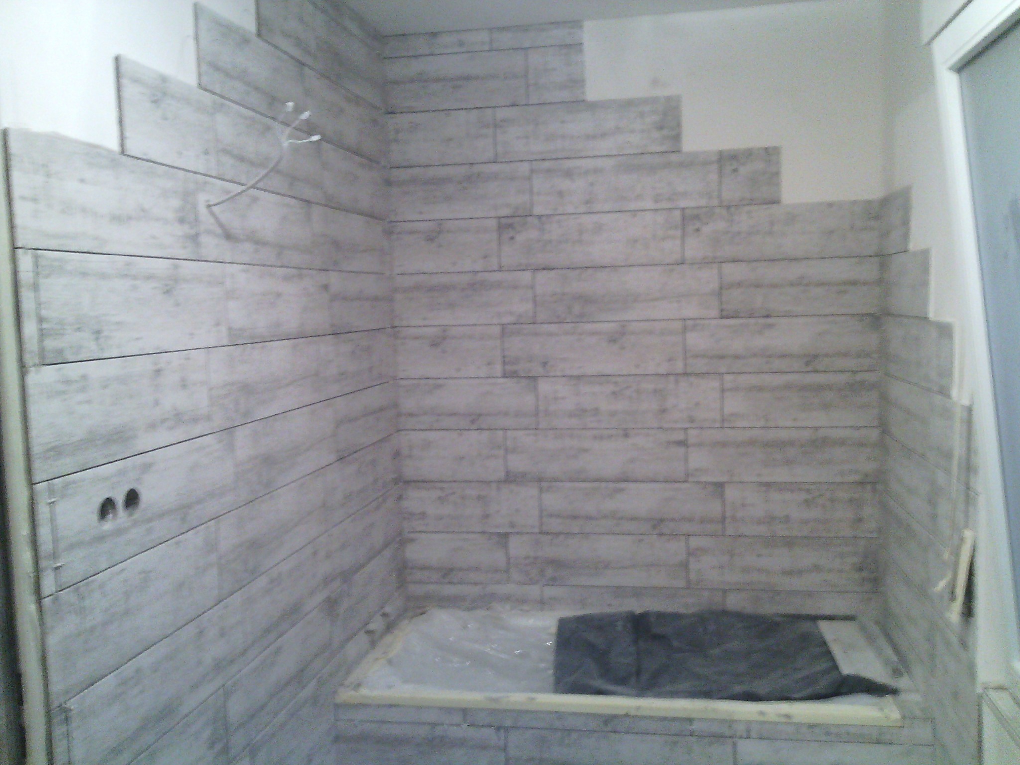 Ściana łazienki w trakcie wykańczania z szarymi płytkami imitującymi drewno, widoczne otwory na gniazdka i wystające kable, zabezpieczona folią podłoga.