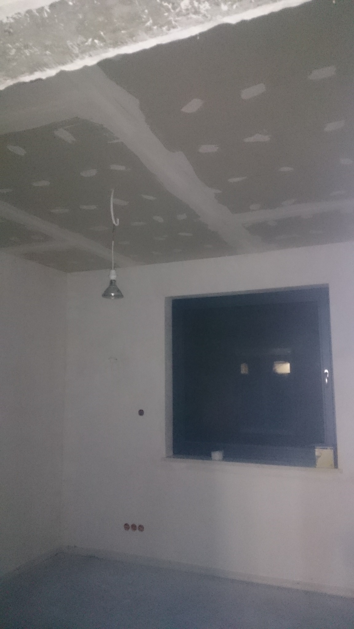 Pokój w trakcie remontu: sufit z płyt gipsowo-kartonowych ze szpachlowanymi łączeniami, wisząca żarówka na kablu, okno i gładkie ściany przygotowane do malowania.