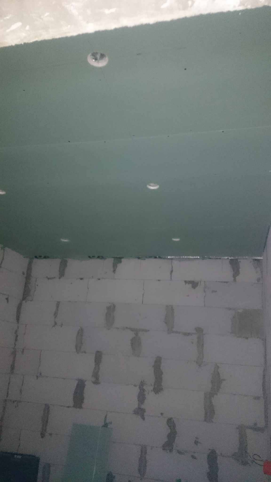 Montaż sufitu podwieszanego z płyt gipsowo-kartonowych z wyciętymi otworami na oświetlenie, widoczna ściana z bloczków betonowych przed wykończeniem.