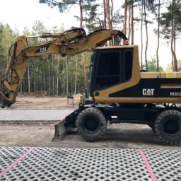 Żółto-czarna koparka CAT M312 z hydraulicznym chwytakiem do rozbiórki, stojąca na placu wyłożonym ażurowymi płytami betonowymi, widok z boku, w tle las.