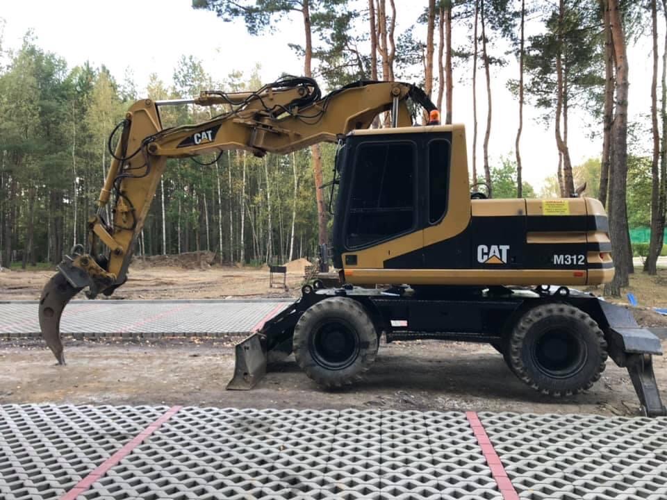 Żółto-czarna koparka CAT M312 z hydraulicznym chwytakiem do rozbiórki, stojąca na placu wyłożonym ażurowymi płytami betonowymi, widok z boku, w tle las.
