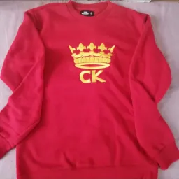 Czerwona bluza dresowa z haftowanym złotym motywem korony i liter CK na środku, leżąca na jasnym tle.