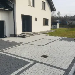 Nowoczesny podjazd z jasnoszarej kostki brukowej z ciemnoszarymi obramowaniami, studzienka kanalizacyjna na środku, przed białym domem z szarą stolarką i garażem.