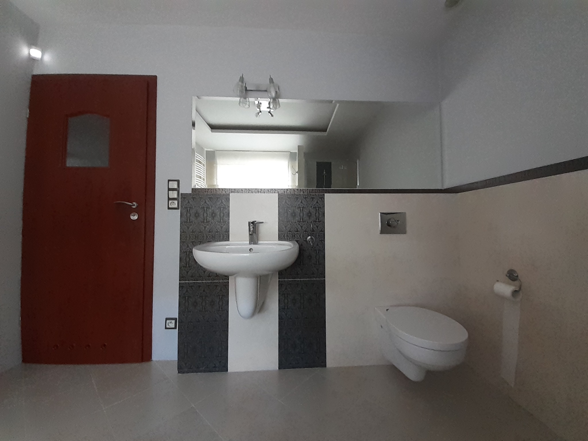 Umywalka z postumentem, wisząca toaleta i lustro w wyremontowanej łazience z szaro-białymi płytkami i brązowymi drzwiami.