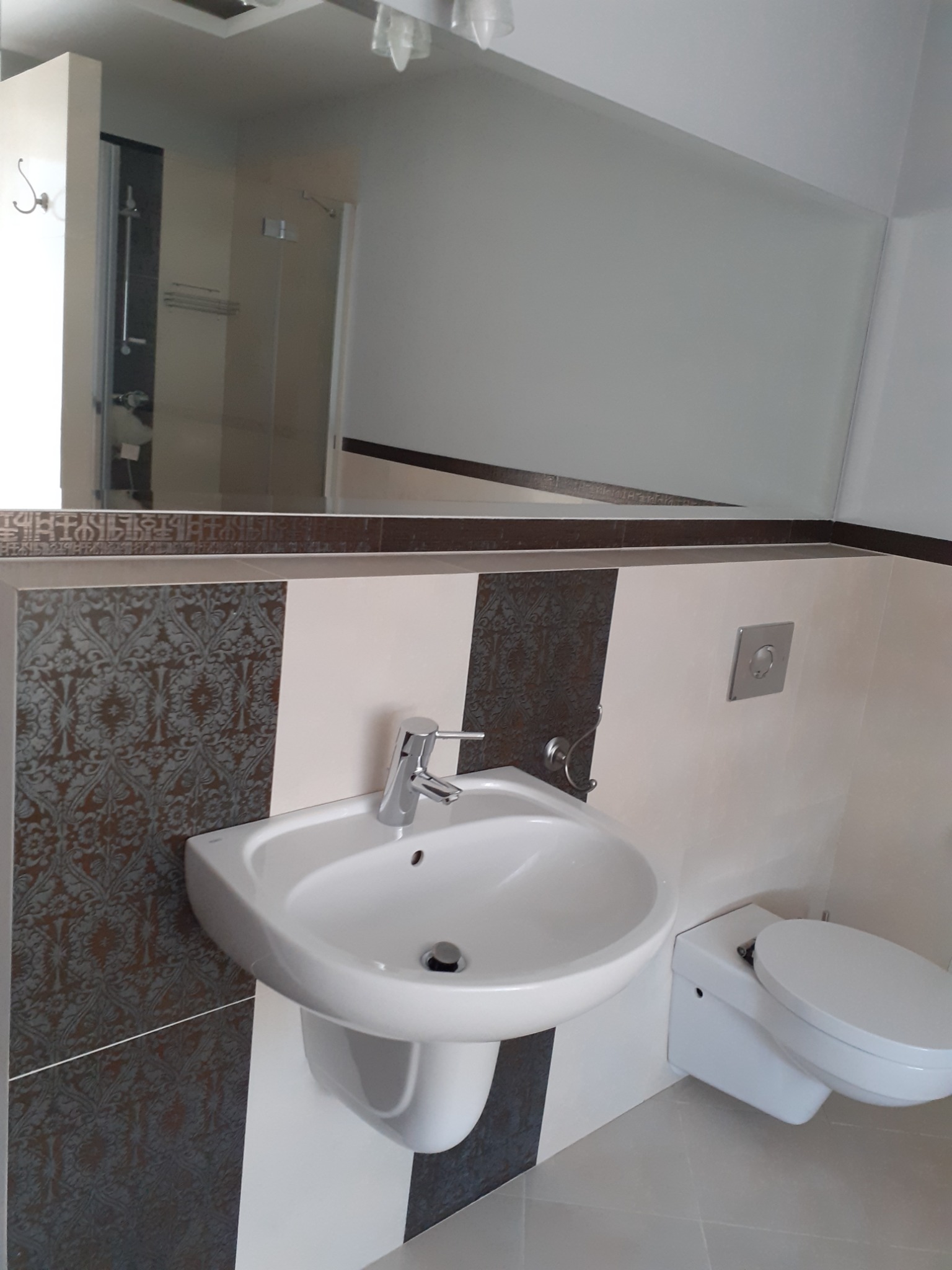 Nowoczesna łazienka z umywalką nablatową, toaletą podwieszaną i dużym lustrem, wykończona płytkami w geometryczne wzory i odcieniach bieli oraz szarości.