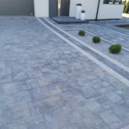 Szara kostka brukowa ułożona przed nowoczesnym domem z szarą stolarką okienną i drzwiową, obok wzdłuż podjazdu pas z jasnych kamieni i krzewów.