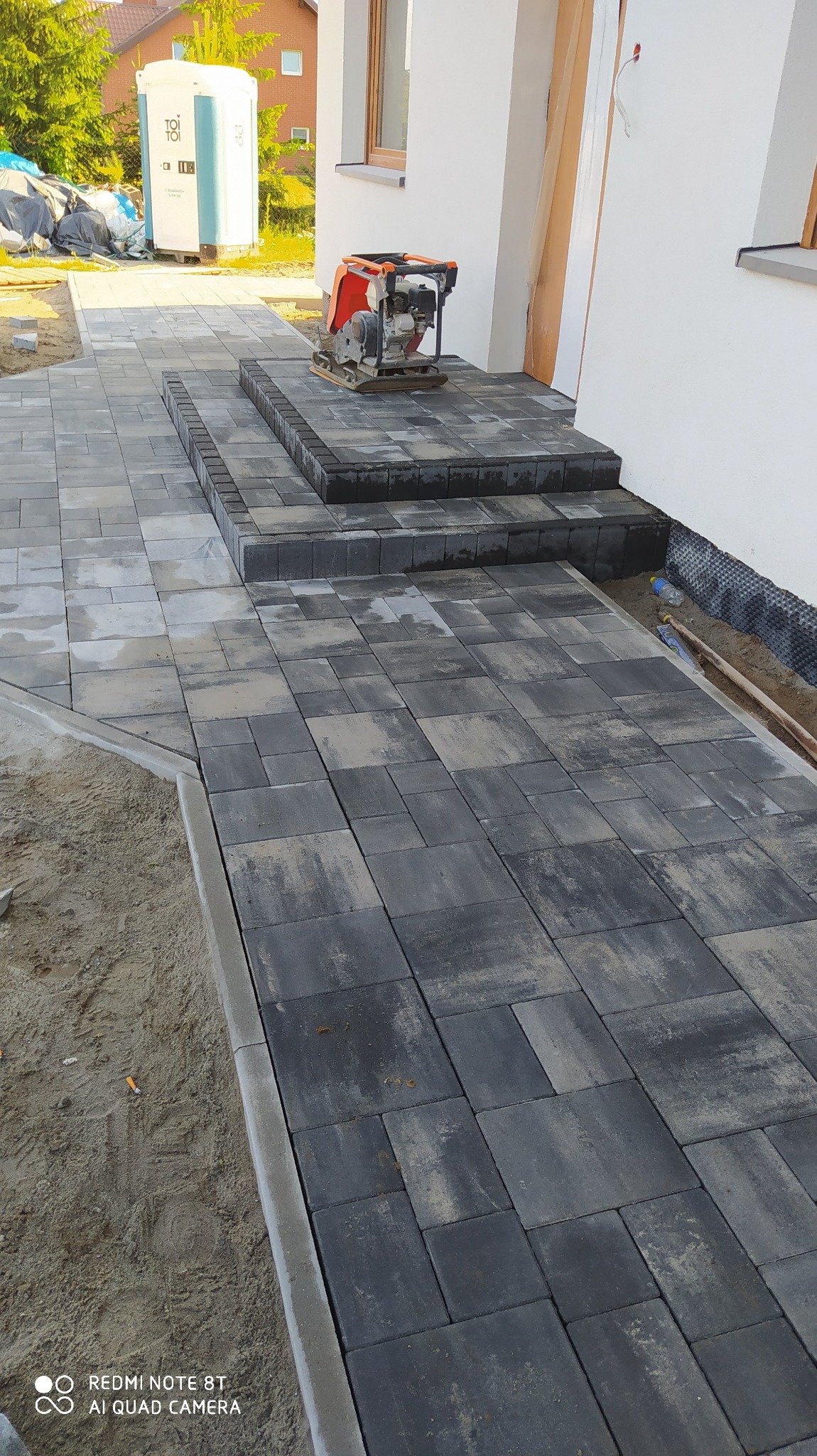 Świeżo ułożona, grafitowa kostka brukowa prowadząca do wejścia domu, z zagęszczarką wibracyjną na schodach i przenośną toaletą w tle, widoczne ślady piasku i niedokończone krawędzie.