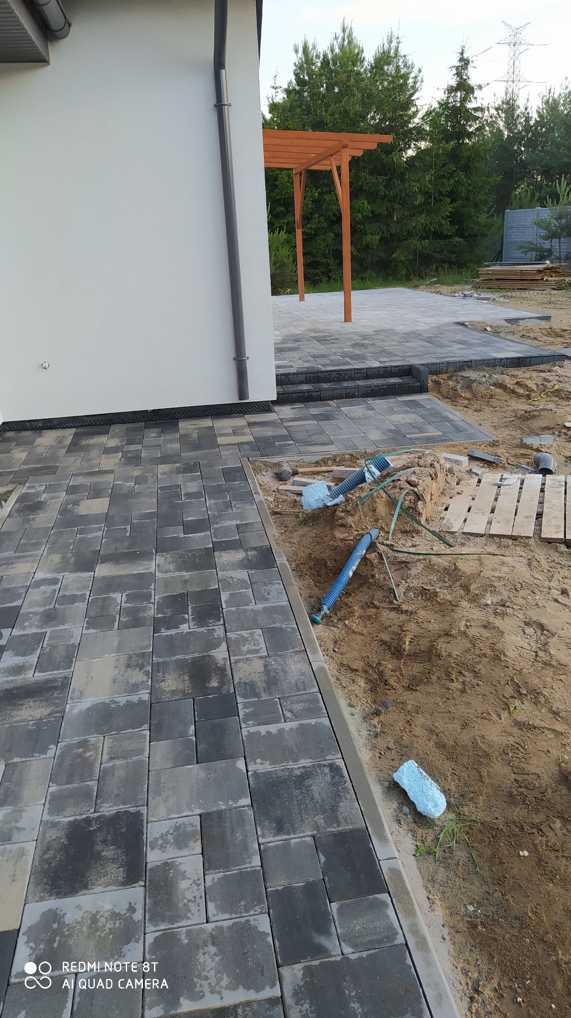 Ułożona kostka brukowa w odcieniach szarości, obok wykop z wystającymi rurami i drewnianymi deskami, pergola w tle.