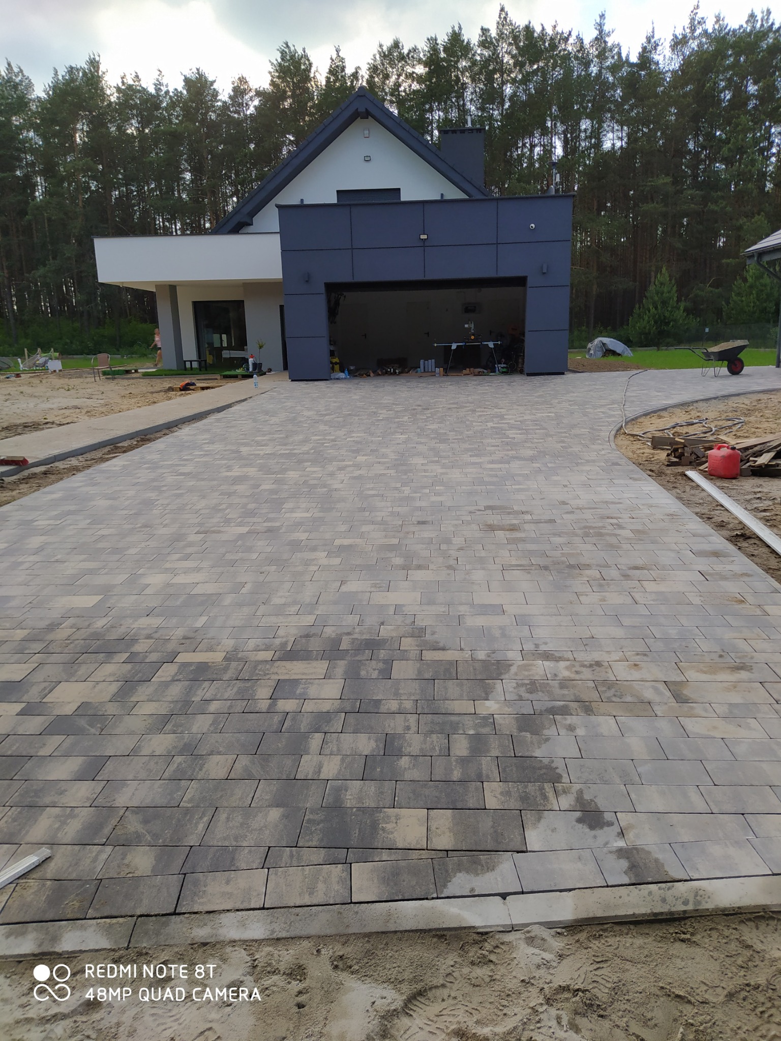 Ułożona kostka brukowa w odcieniach szarości i beżu prowadząca do nowoczesnego domu z garażem, widoczny piasek i elementy wykończeniowe.