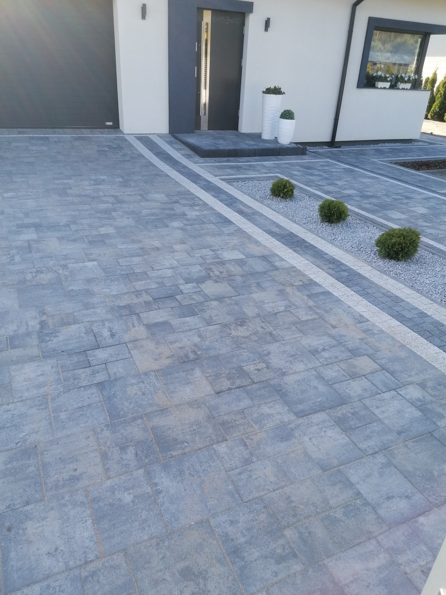 Szara kostka brukowa ułożona przed nowoczesnym domem z szarą stolarką okienną i drzwiową, obok wzdłuż podjazdu pas z jasnych kamieni i krzewów.