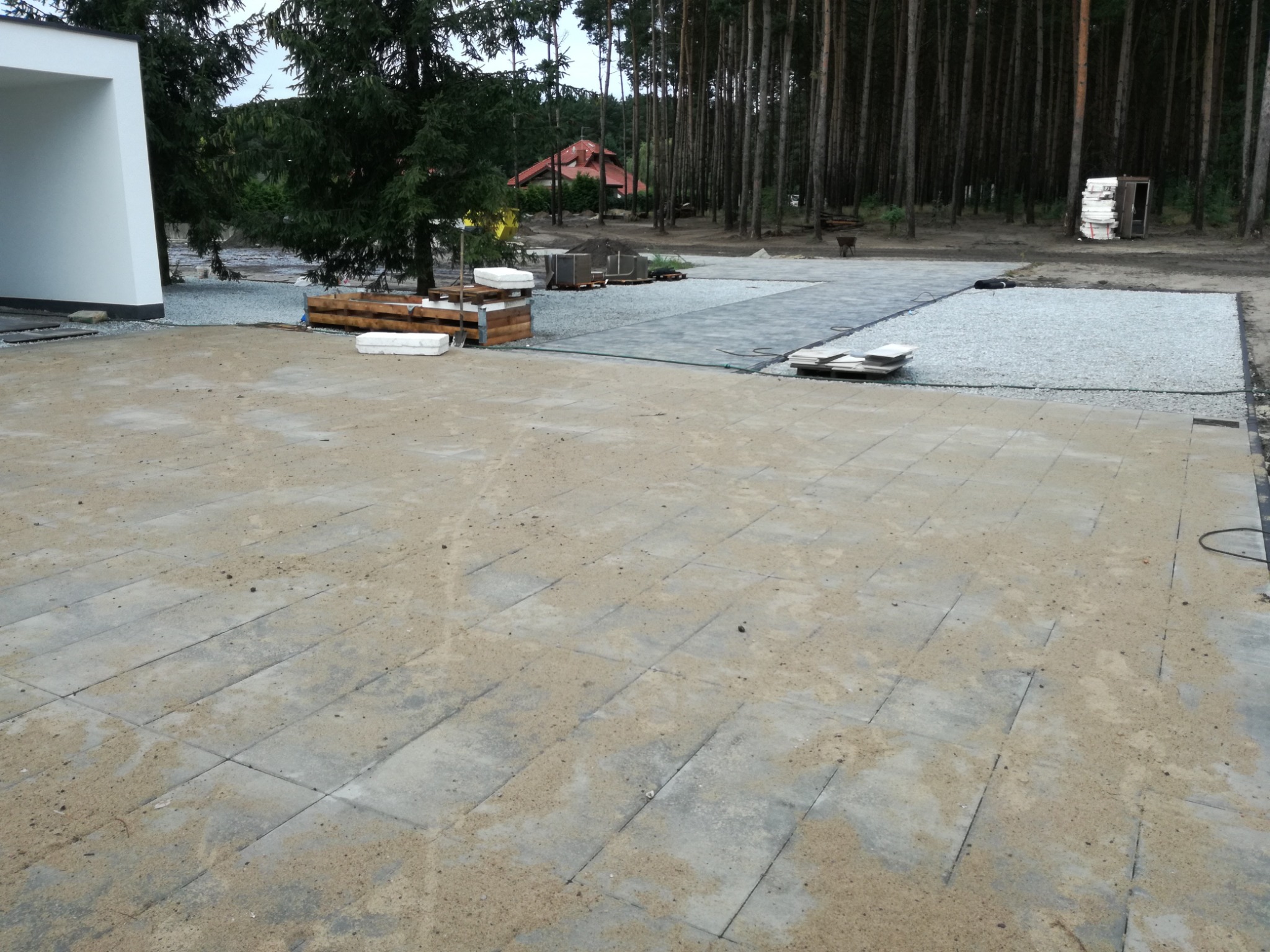 Fragment układanej nawierzchni z kostki brukowej, widoczne przygotowane podłoże z grysu, w tle budynek z białymi ścianami i las sosnowy.