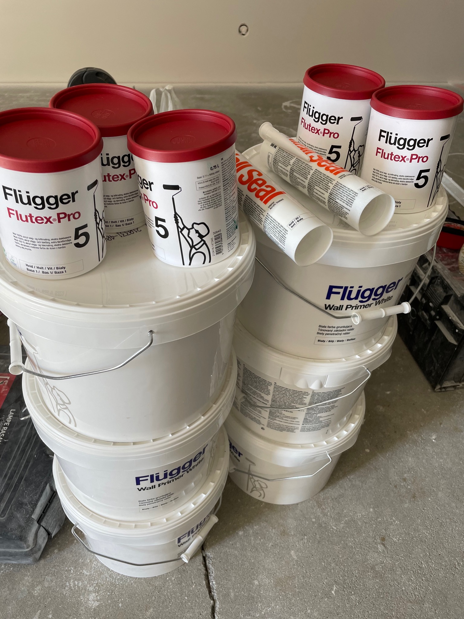 Stos białych wiader z farbami i gruntami marki Flugger, z czerwonymi pokrywkami na górze, oraz tuby z akrylem, ustawione na szarej podłodze w trakcie remontu.