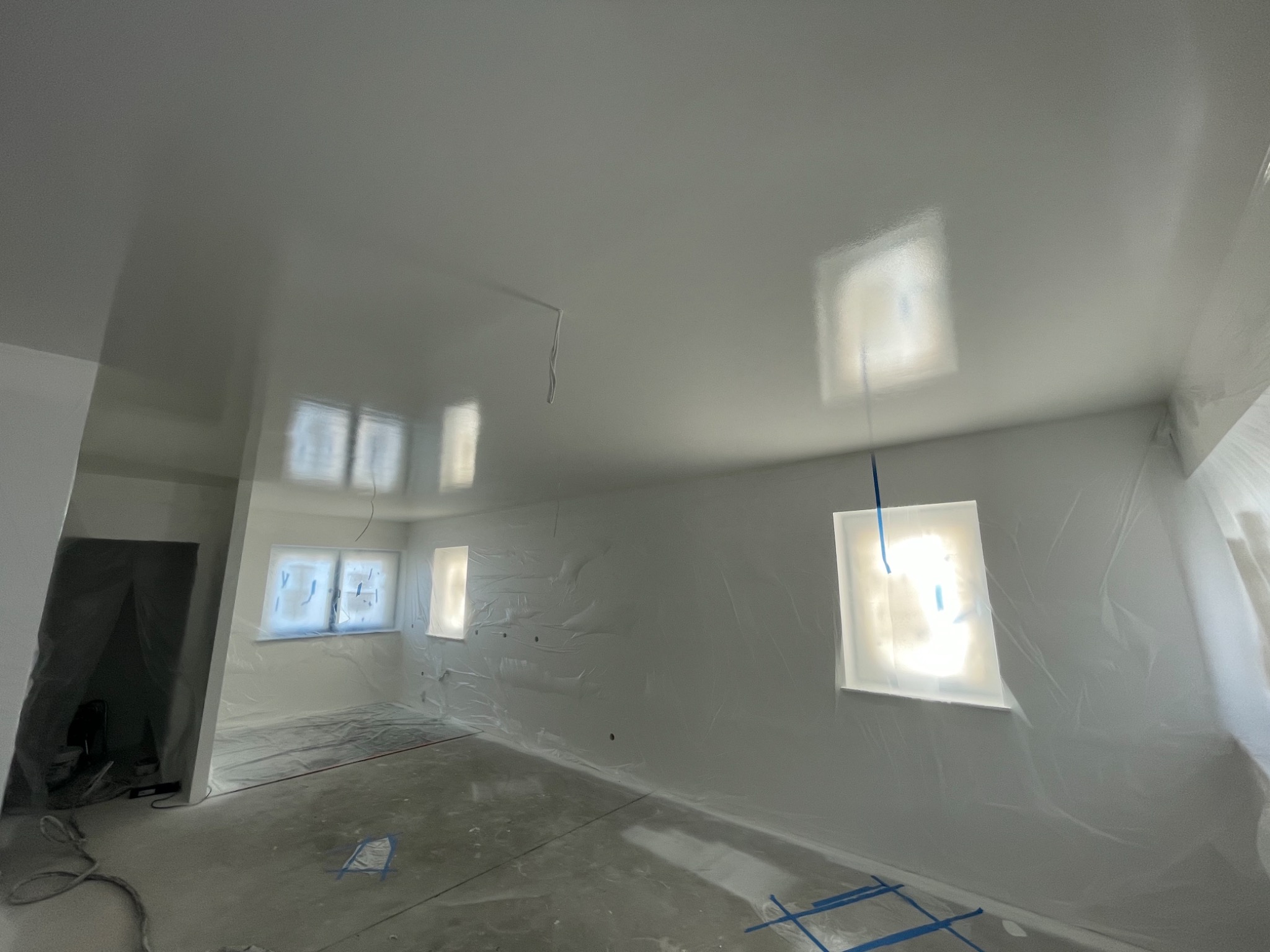 Pokój przygotowany do malowania: okna i ściany zabezpieczone folią, podłoga zakryta, świeżo położona biała warstwa na ścianach i suficie, widoczne ślady taśmy malarskiej.