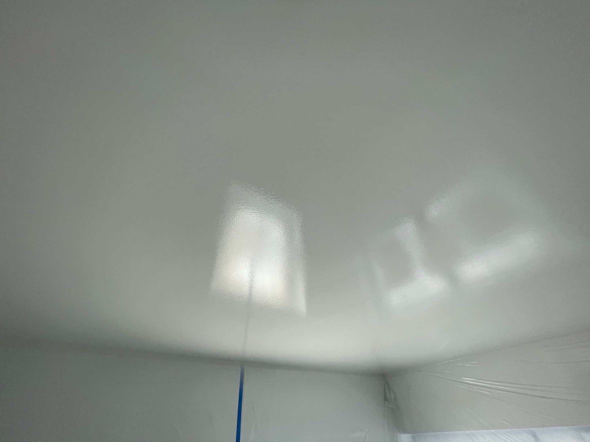 Świeżo pomalowany biały sufit z odbiciami światła, zabezpieczony folią narożnik ściany z widocznym niebieskim paskiem taśmy malarskiej.