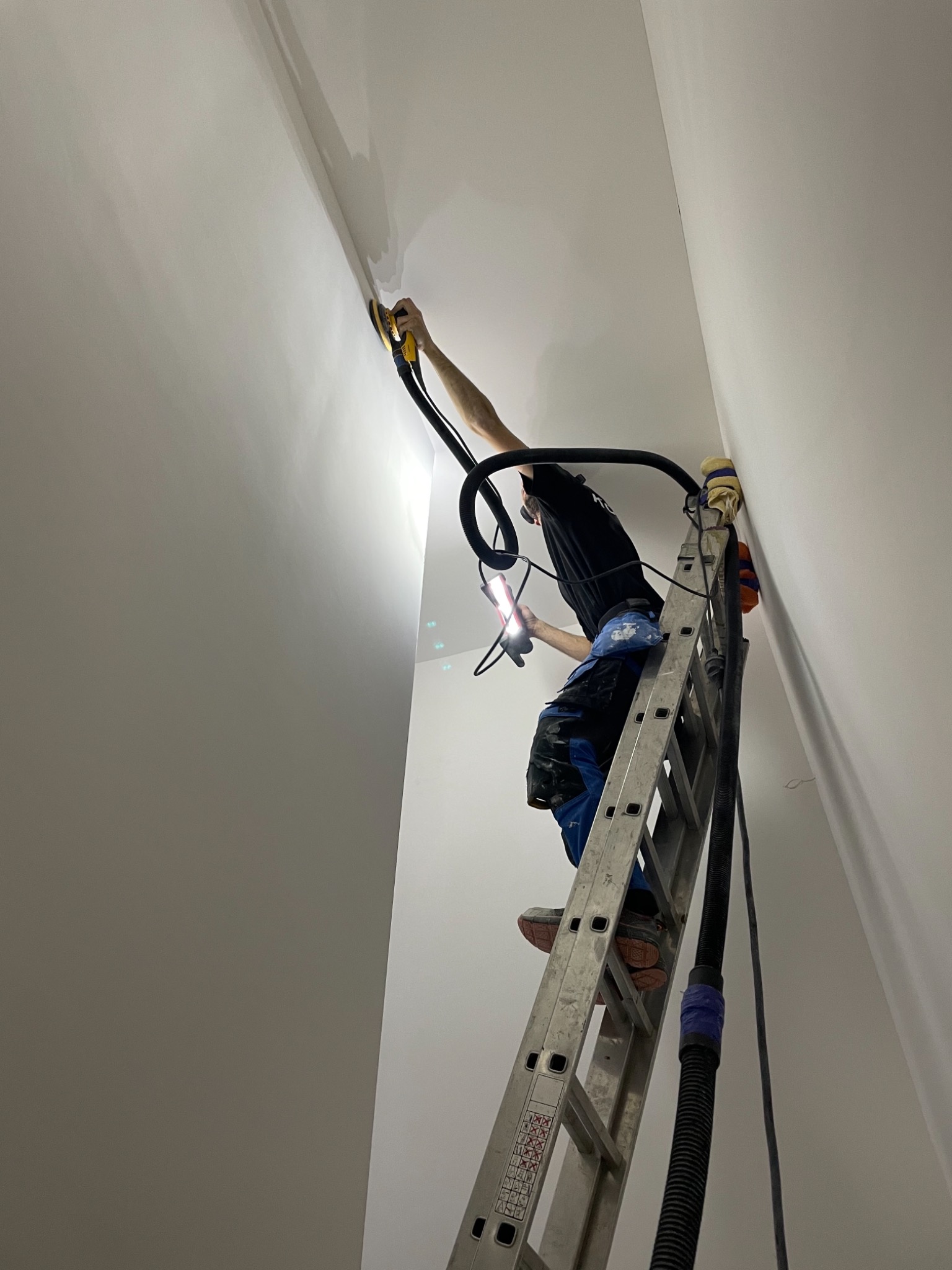 Pracownik na drabinie szlifuje łączenie ścian i sufitu za pomocą szlifierki z odkurzaczem, oświetlając sobie miejsce pracy latarką.