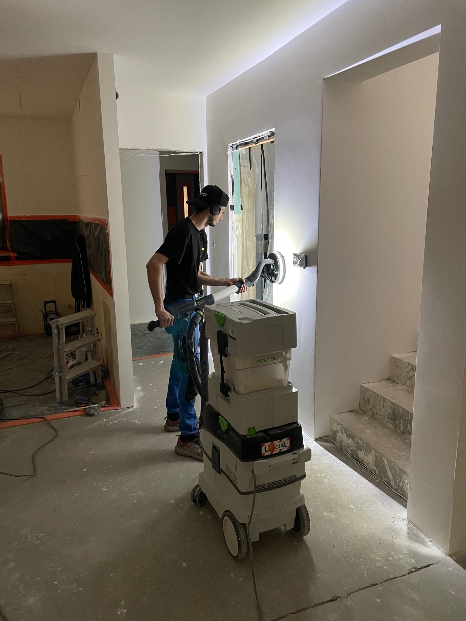 Pracownik w trakcie szlifowania ściany wewnątrz budynku za pomocą szlifierki z odkurzaczem przemysłowym Festool, widoczne przygotowane do malowania ściany i schody w surowym stanie.