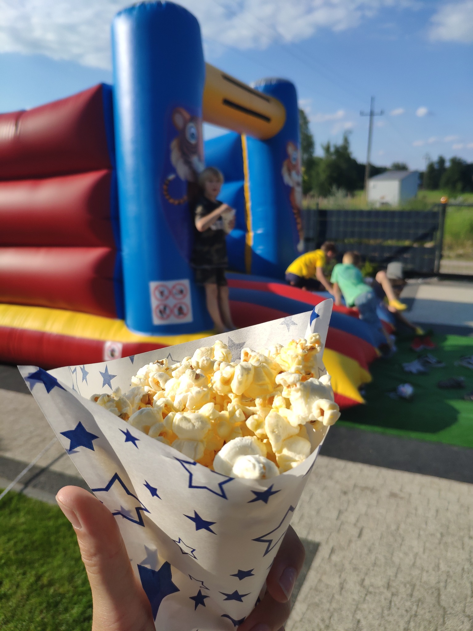 Popcorn w papierowym rożku z gwiazdkami, w tle dmuchany zamek i bawiące się dzieci.