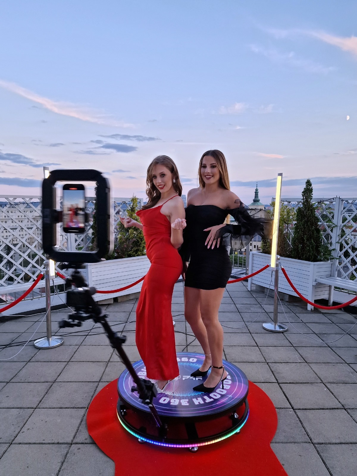 Dwie kobiety w eleganckich sukienkach pozują na platformie fotobudki 360 z podświetleniem LED na tle miejskiego krajobrazu o zmierzchu.
