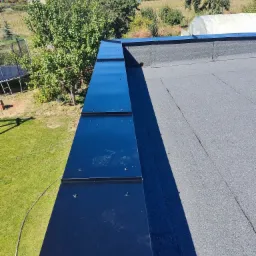 Widok z góry na fragment dachu pokrytego papą i obróbki blacharskie w kolorze czarnym, z widocznym ogrodem i zabudowaniami w tle w słoneczny dzień.