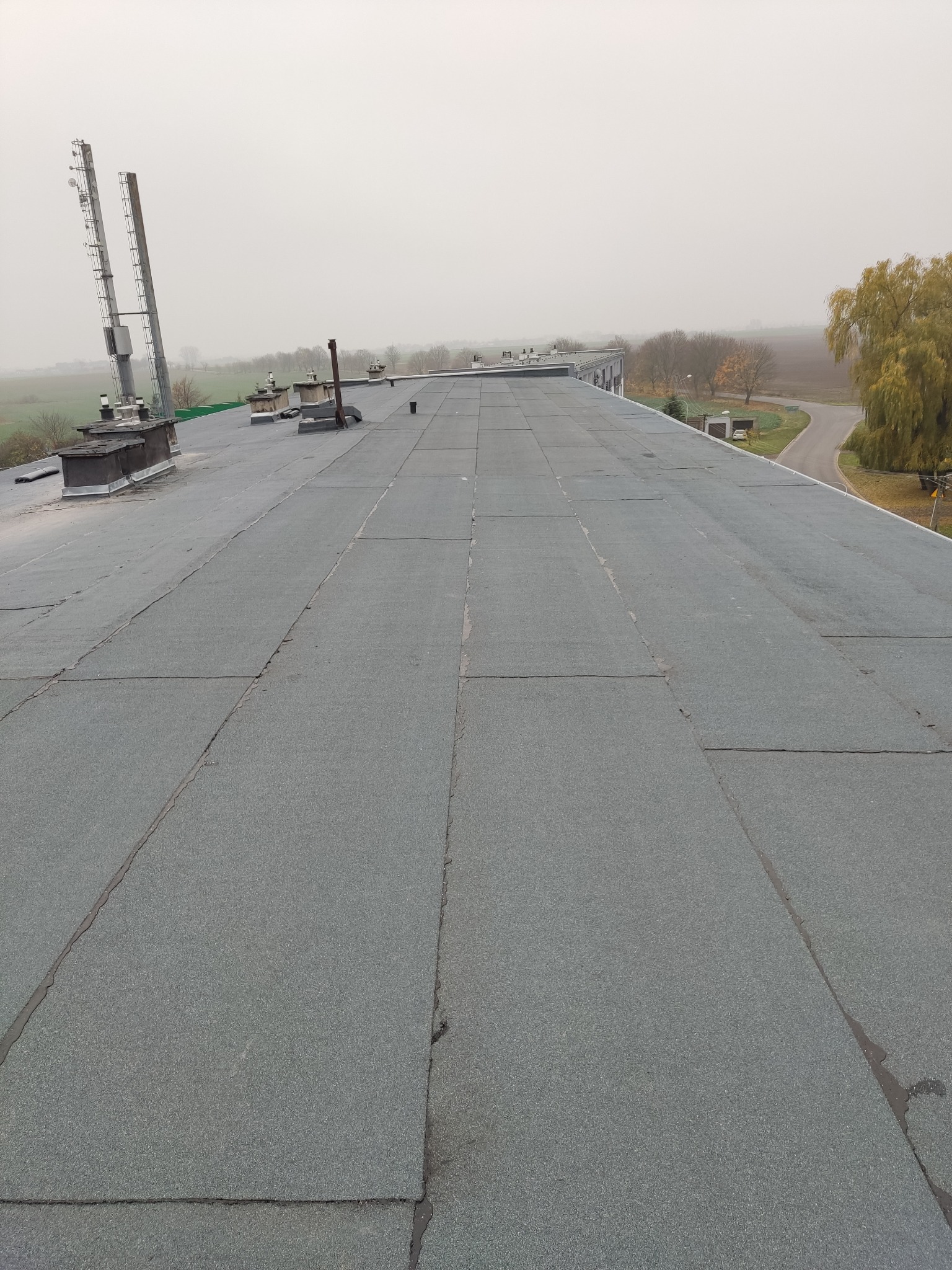 Płaski dach pokryty papą termozgrzewalną w szarym kolorze, widoczne kominy wentylacyjne i maszty antenowe, w tle zarys krajobrazu w pochmurny dzień.