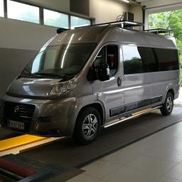 Auto-Komplex Michał Zielonka - Szary van na stanowisku diagnostycznym z rolkami do badania hamulców, widok z przodu lekko z boku, z zamontowanym bagażnikiem dachowym.