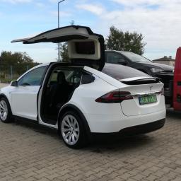 Auto-Komplex Michał Zielonka - Biała Tesla Model X z otwartymi drzwiami typu Falcon Wing, zaparkowana na kostce brukowej obok czerwonego samochodu dostawczego i czarnego SUV-a.