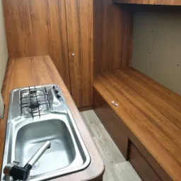 Wnętrze kampera z zabudową meblową w odcieniach brązu, z kuchenką i zlewem po lewej stronie oraz miejscem do spania po prawej, wykonane w Lyski.