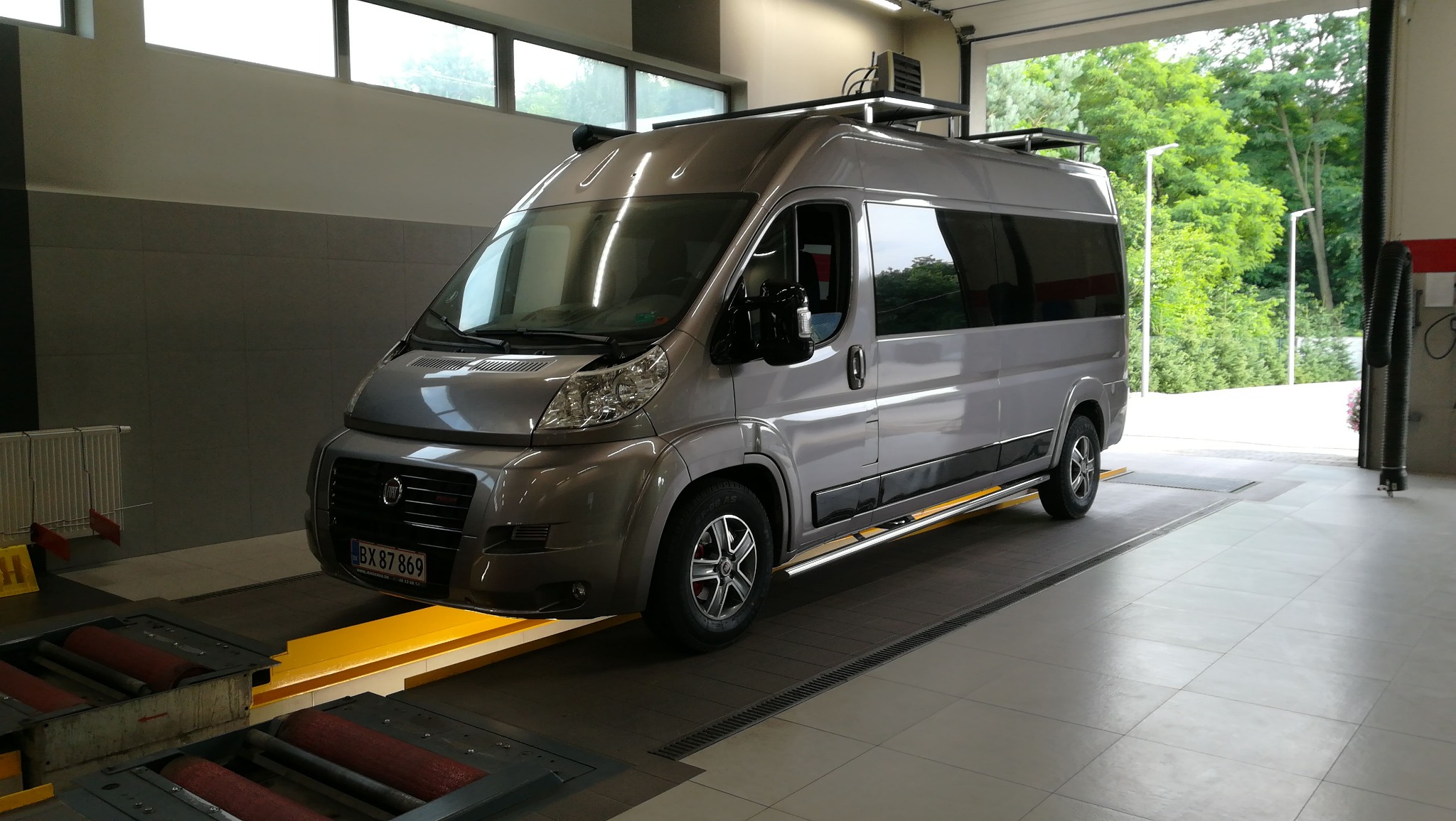 Szary van na stanowisku diagnostycznym z rolkami do badania hamulców, widok z przodu lekko z boku, z zamontowanym bagażnikiem dachowym.