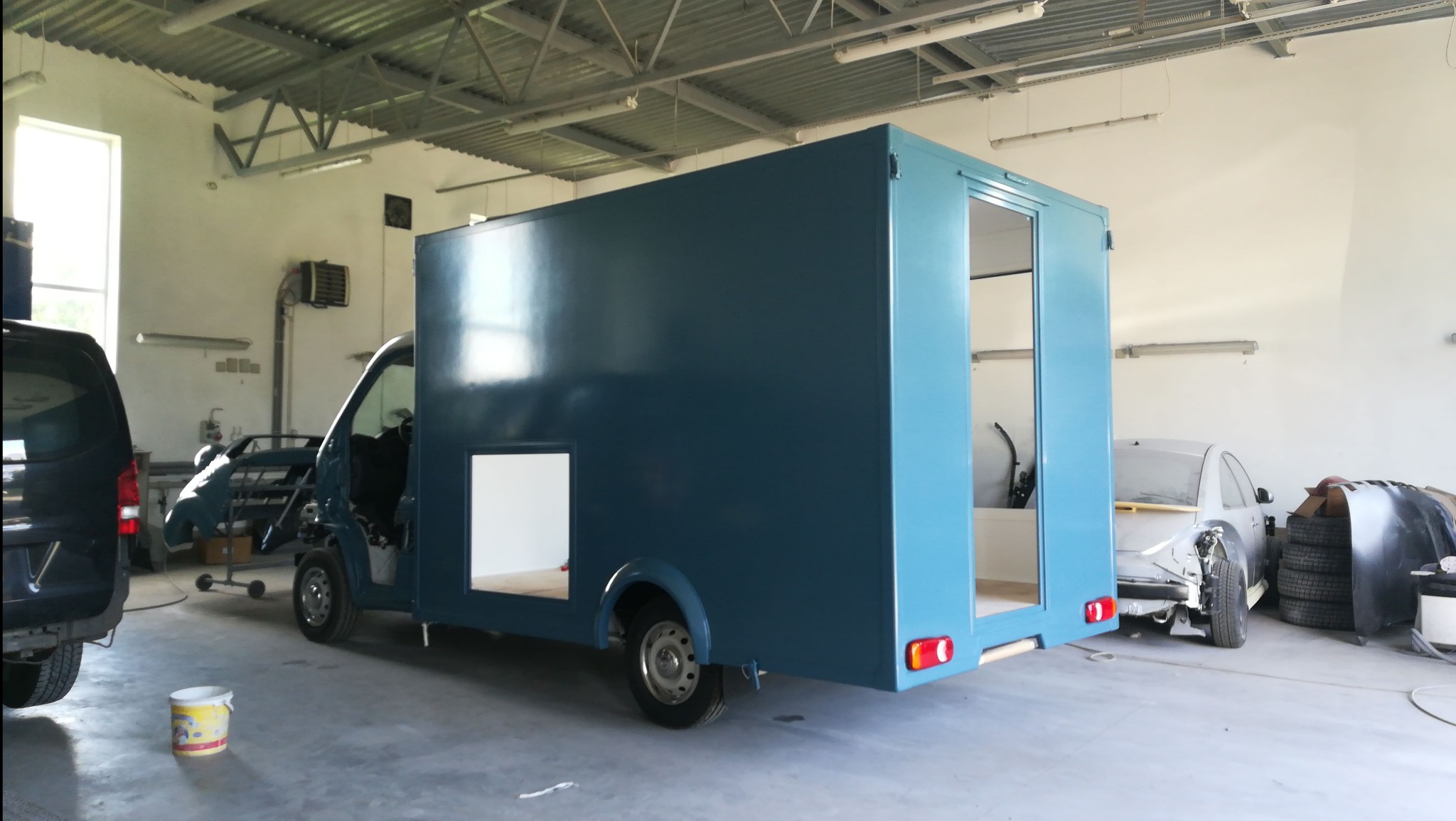 Niebieski samochód dostawczy w trakcie lakierowania w warsztacie, z otwartymi drzwiami i widocznym wnętrzem skrzyni ładunkowej, inne pojazdy w tle.