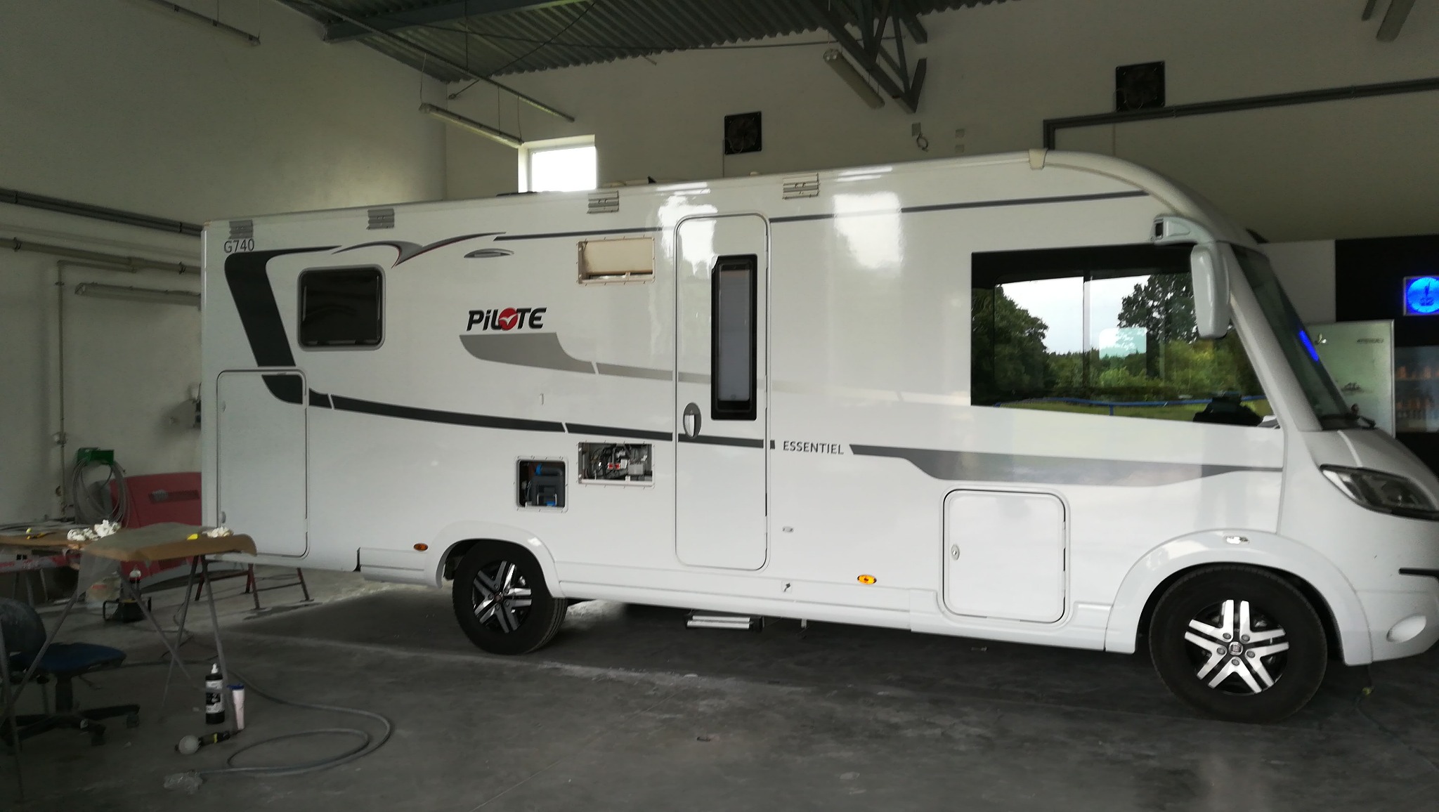 Biały kamper marki Pilote Essentiel G740 w warsztacie, widoczne otwarte drzwiczki serwisowe i stolik roboczy z narzędziami.