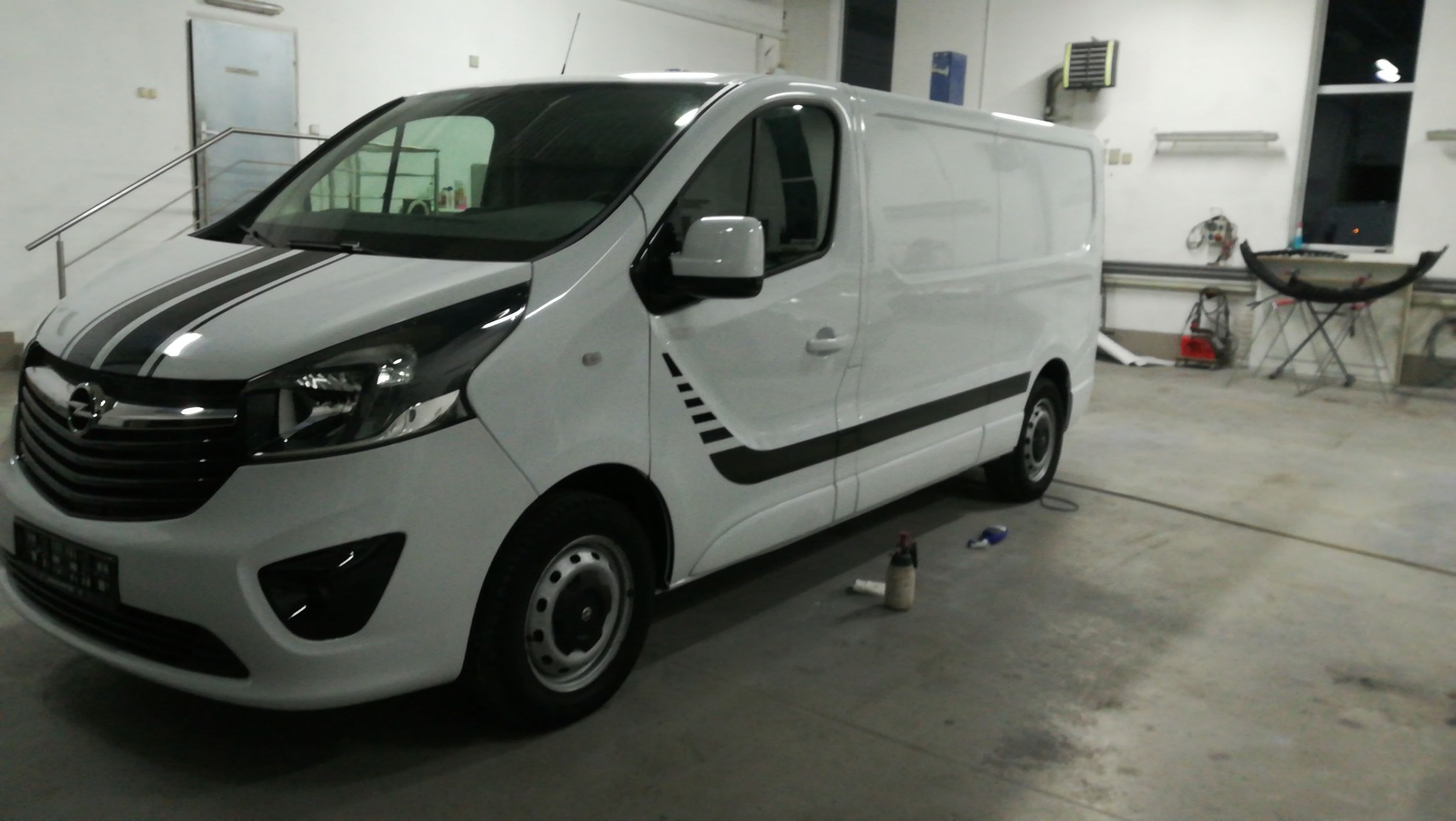 Biały Opel Vivaro z czarnymi pasami dekoracyjnymi na karoserii, widoczny w warsztacie samochodowym.