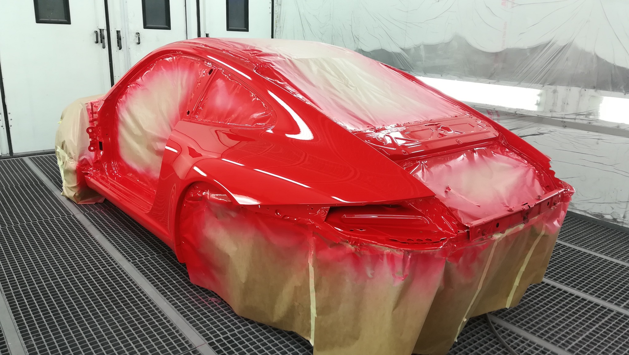 Czerwone nadwozie samochodu sportowego w trakcie lakierowania, częściowo zabezpieczone folią i papierem maskującym, widoczne na podłodze kraty lakiernicze.