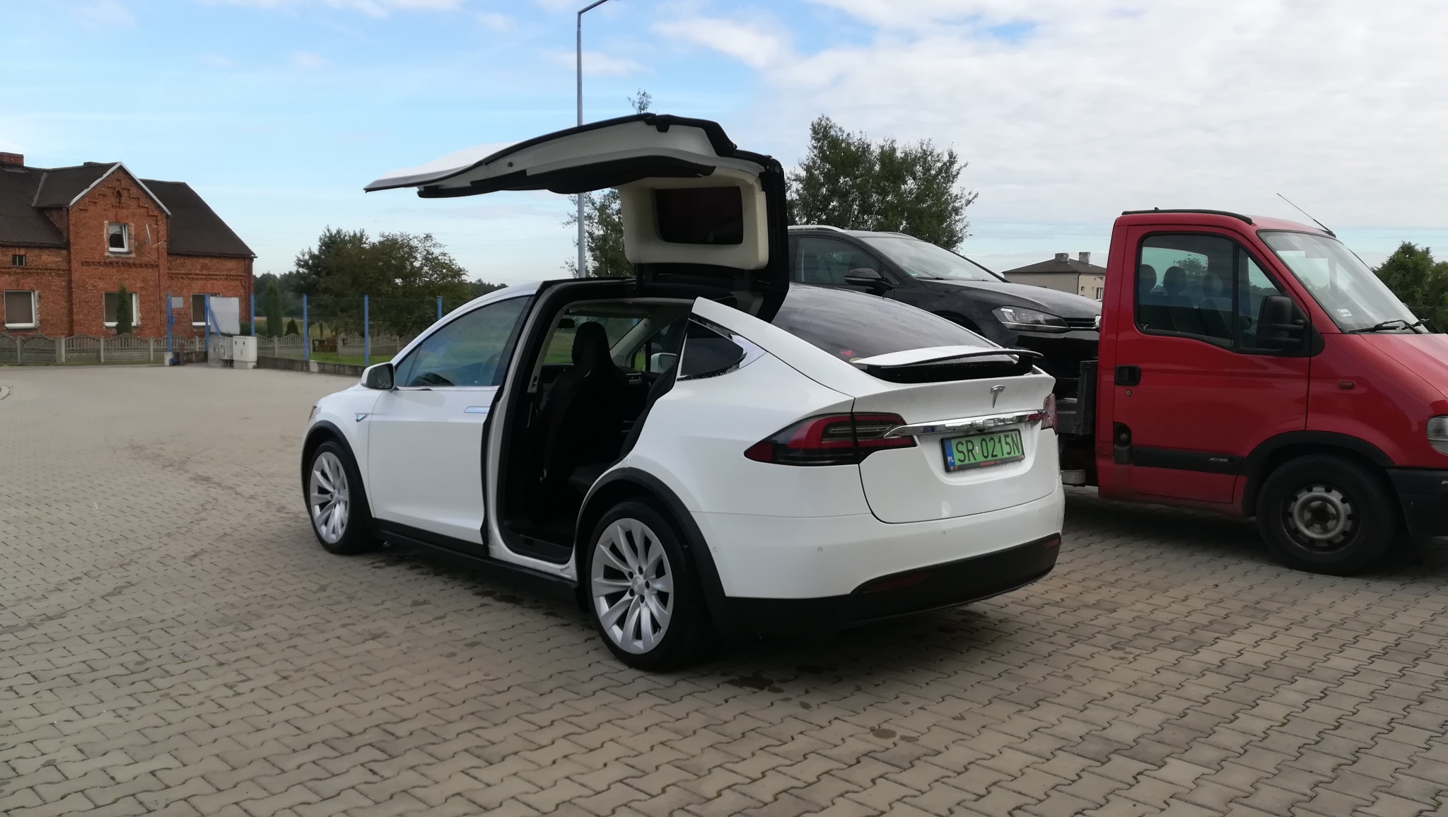 Biała Tesla Model X z otwartymi drzwiami typu Falcon Wing, zaparkowana na kostce brukowej obok czerwonego samochodu dostawczego i czarnego SUV-a.