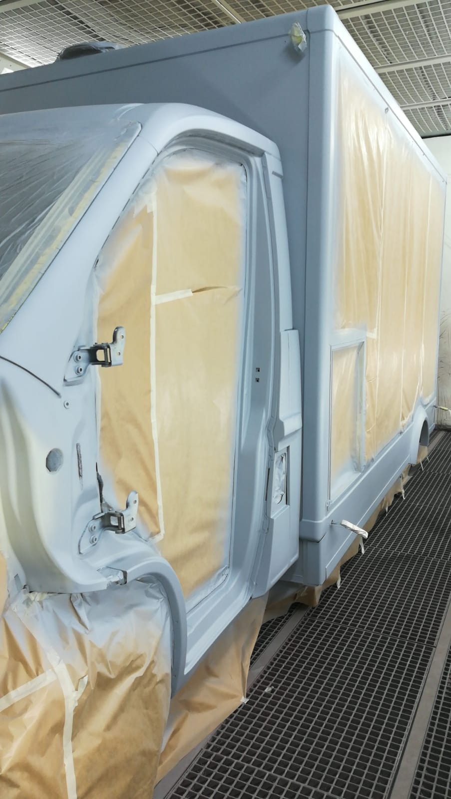 Półciężarówka w trakcie lakierowania w kabinie lakierniczej, zabezpieczona papierem i taśmą malarską, widoczna krata podłogowa.