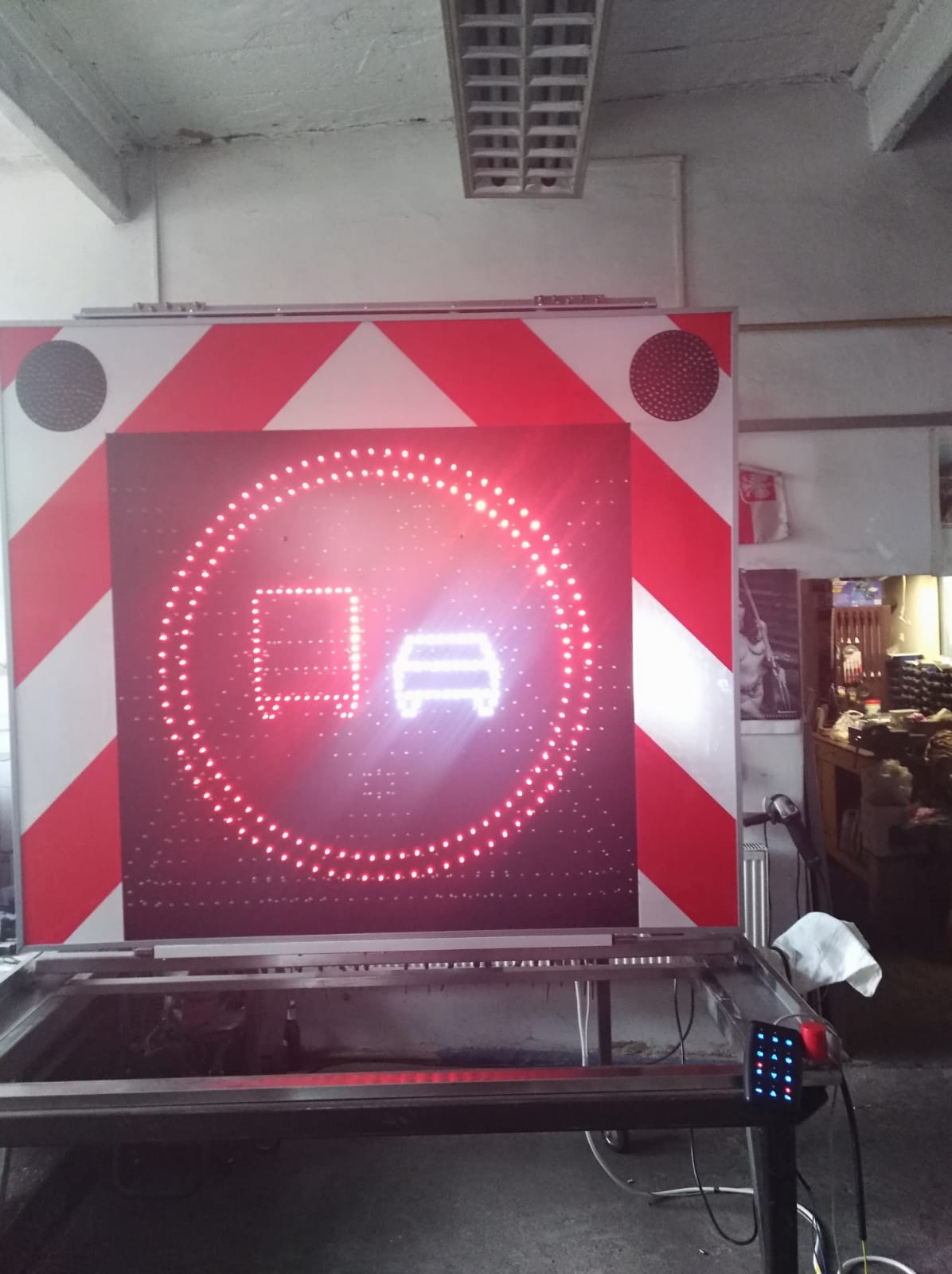 Duży znak drogowy LED z czerwonymi i białymi pasami ostrzegawczymi, wyświetlający ikonę samochodu osobowego i ciężarowego w czerwonym okręgu, prawdopodobnie zakaz wjazdu, w warsztacie.