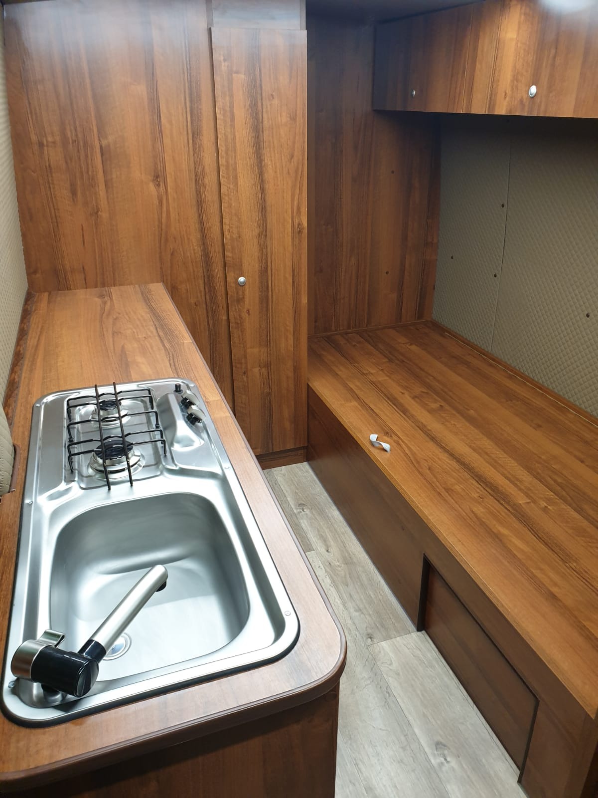 Wnętrze kampera z zabudową meblową w odcieniach brązu, z kuchenką i zlewem po lewej stronie oraz miejscem do spania po prawej, wykonane w Lyski.