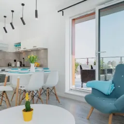 Jasne wnętrze mieszkania z aneksem kuchennym, stołem z krzesłami, balkonem i turkusowym fotelem z kocykiem