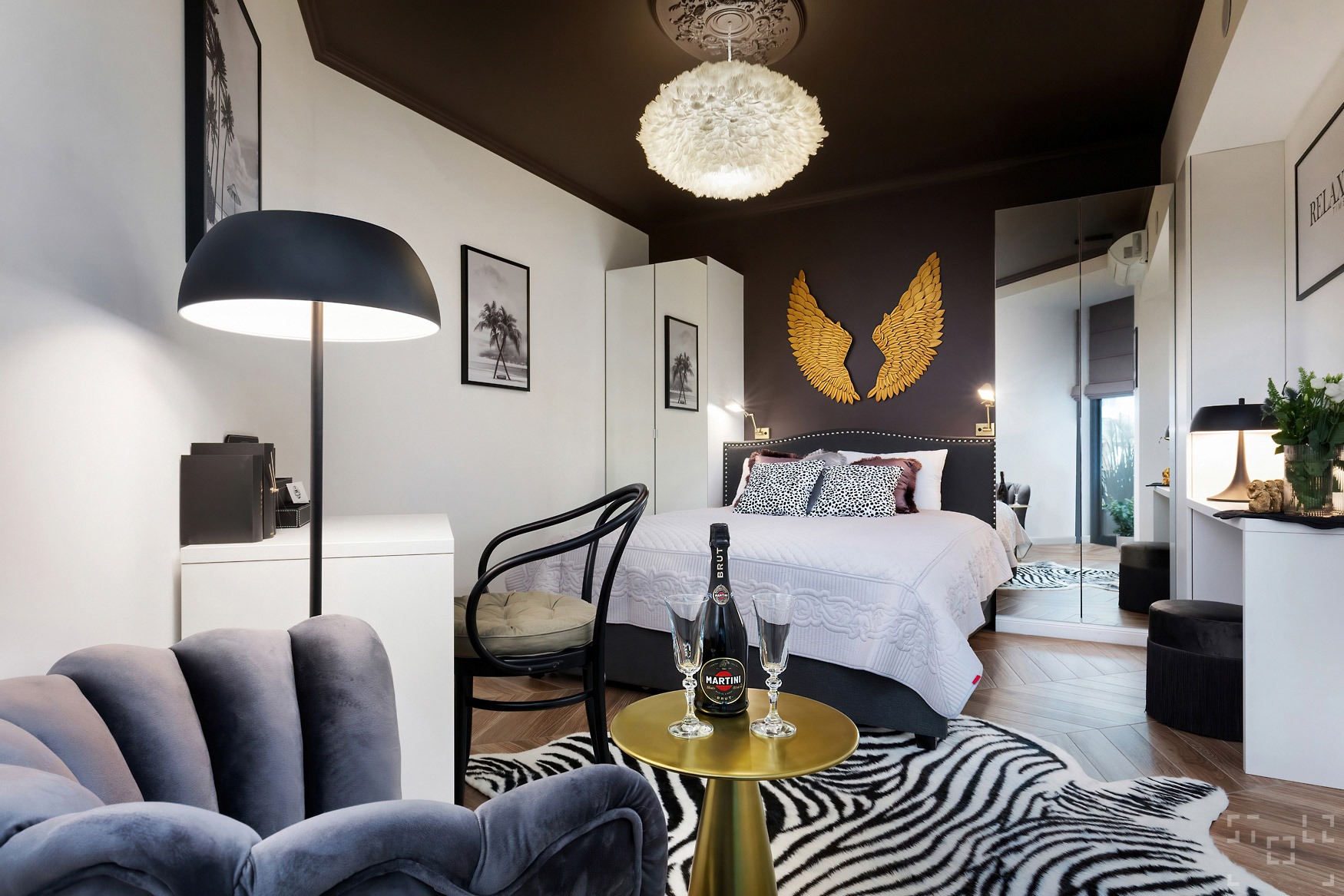 Elegancka sypialnia z łóżkiem, złotymi skrzydłami na ścianie, lampą z piór, stolikiem z butelką Martini i dywanem w zebrę.