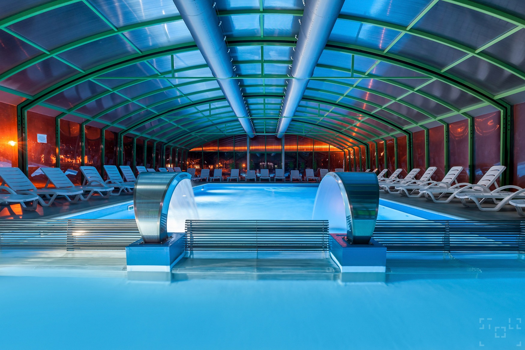 Kryty basen z leżakami, oświetlony niebieskim światłem, z ozdobnymi fontannami i przeszklonym dachem.