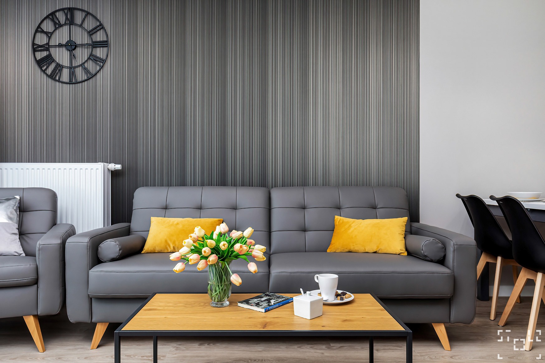 Wnętrze pokoju dziennego z szarą sofą, żółtymi poduszkami, bukietem tulipanów na drewnianym stoliku kawowym, zegarem na ścianie w tle i elementami dekoracyjnymi.