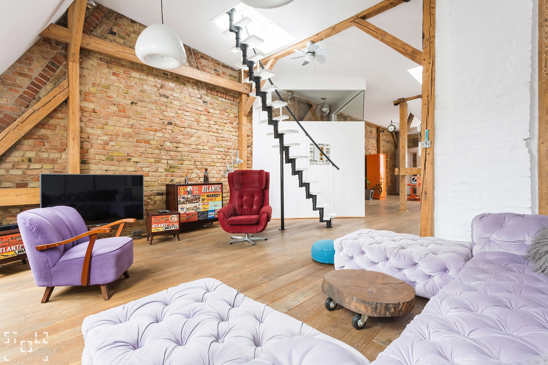 Przestronny salon w stylu loft z ceglaną ścianą, drewnianymi belkami, spiralnymi schodami, fotelem i sofą w odcieniach fioletu, oraz stolikiem kawowym z pnia drzewa na kółkach.
