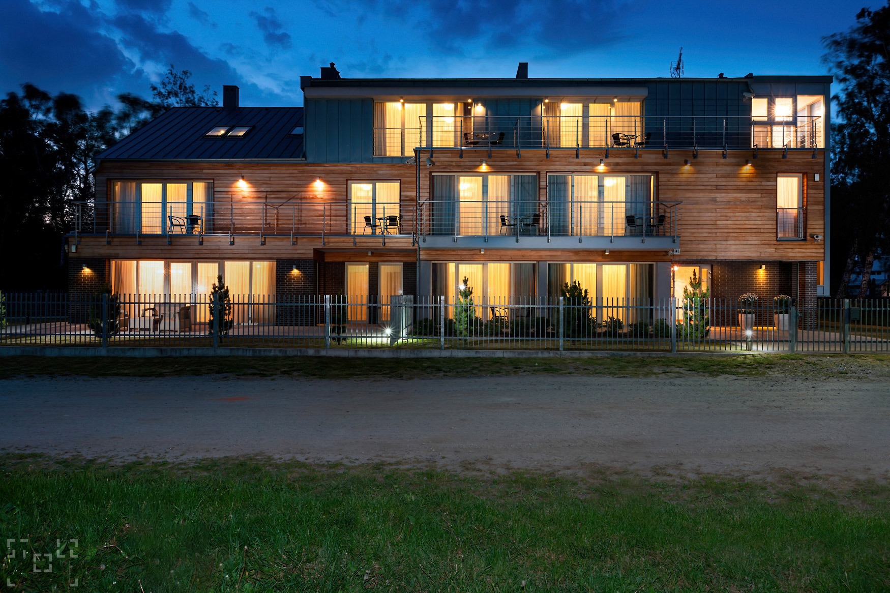 Dwupiętrowy budynek mieszkalny z drewnianą elewacją i balkonami, oświetlony ciepłym światłem w nocy, widziany zza metalowego ogrodzenia.