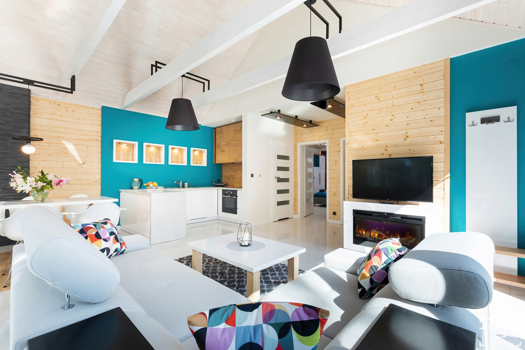 Jasne, przestronne wnętrze domu z drewnianymi panelami, turkusowymi akcentami i nowoczesnym kominkiem elektrycznym pod telewizorem, widok z kanapy na otwartą kuchnię i jadalnię, czarne lampy wiszące.