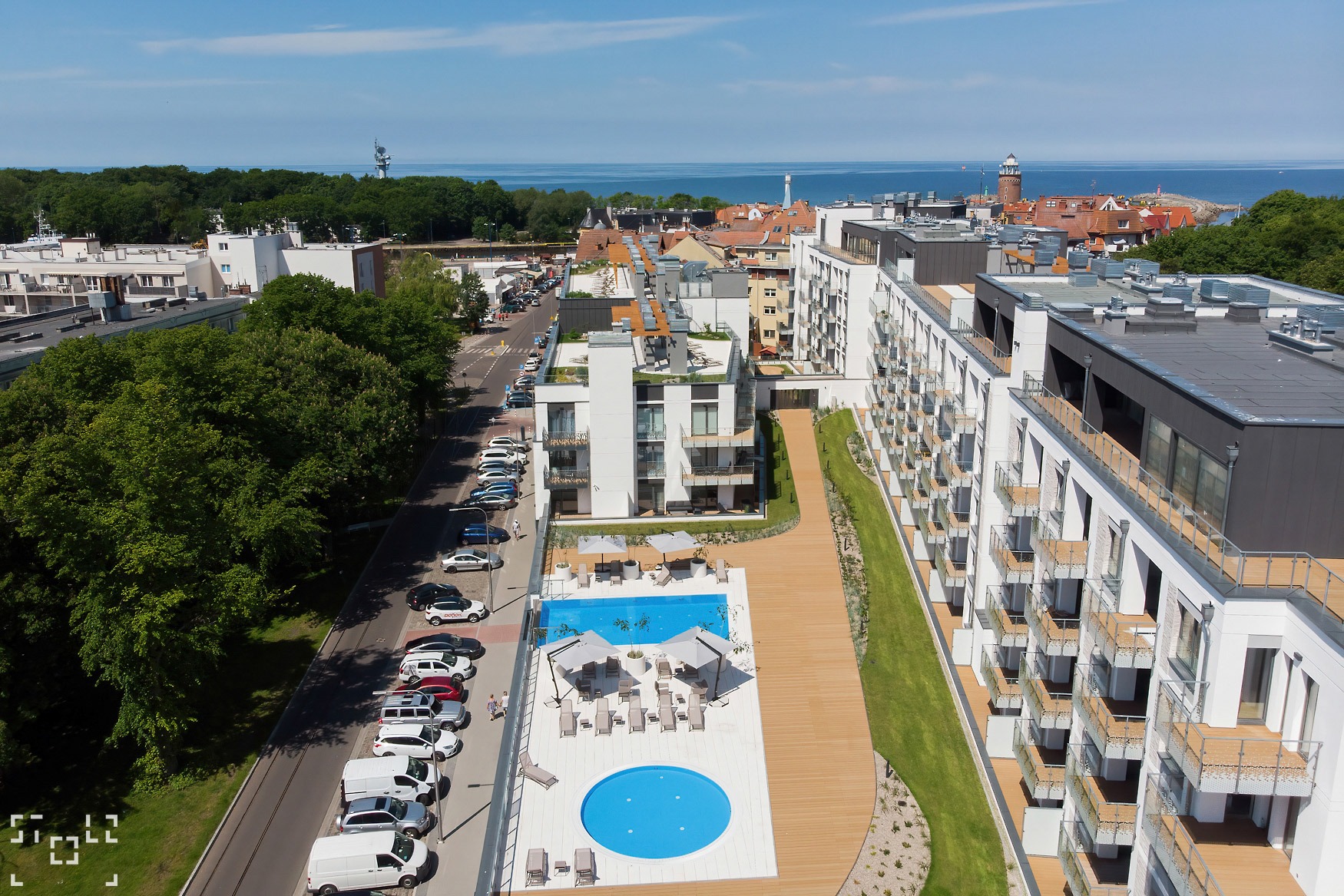 Zdjęcie z drona przedstawia nowoczesny kompleks apartamentów z basenami, drewnianym tarasem, zielonymi skarpami i widokiem na morze w tle.