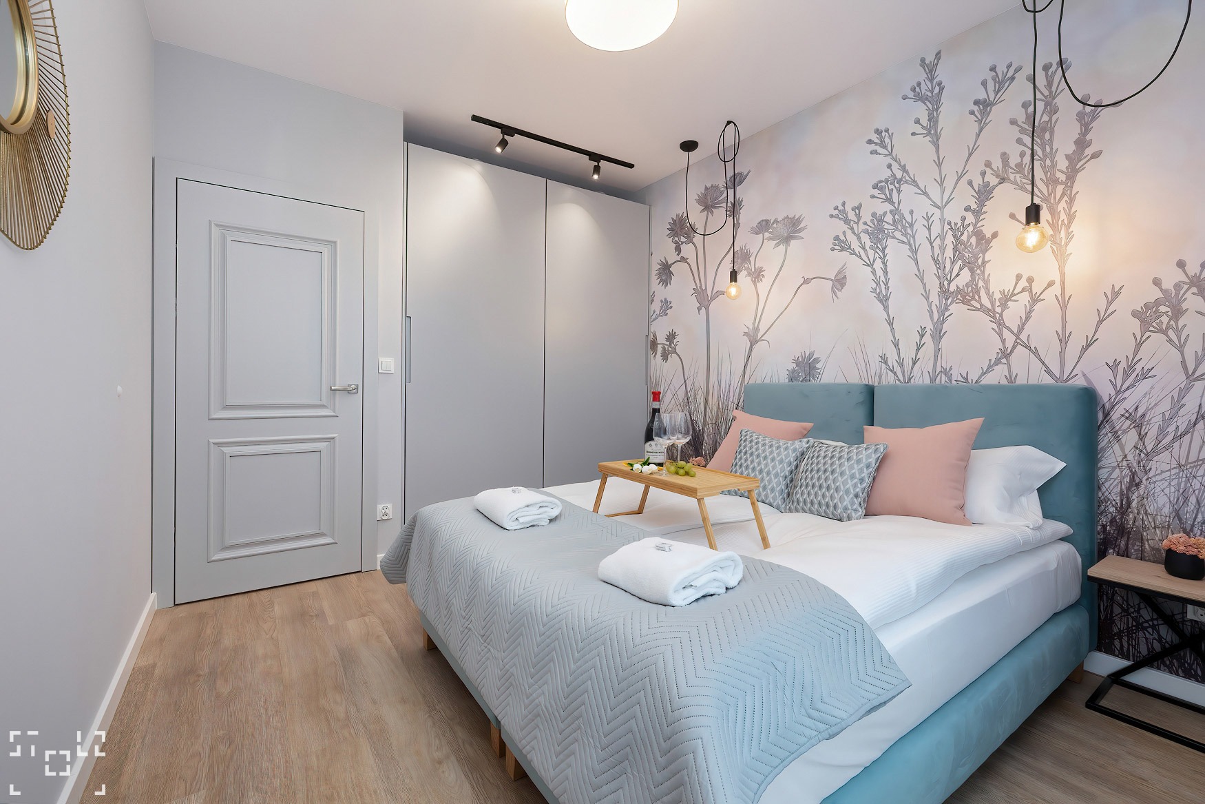 Jasna sypialnia z łóżkiem, na którym leży taca z winem i owocami, oraz dekoracyjną tapetą w motywy roślinne, oświetlona lampami wiszącymi i szynoprzewodami.