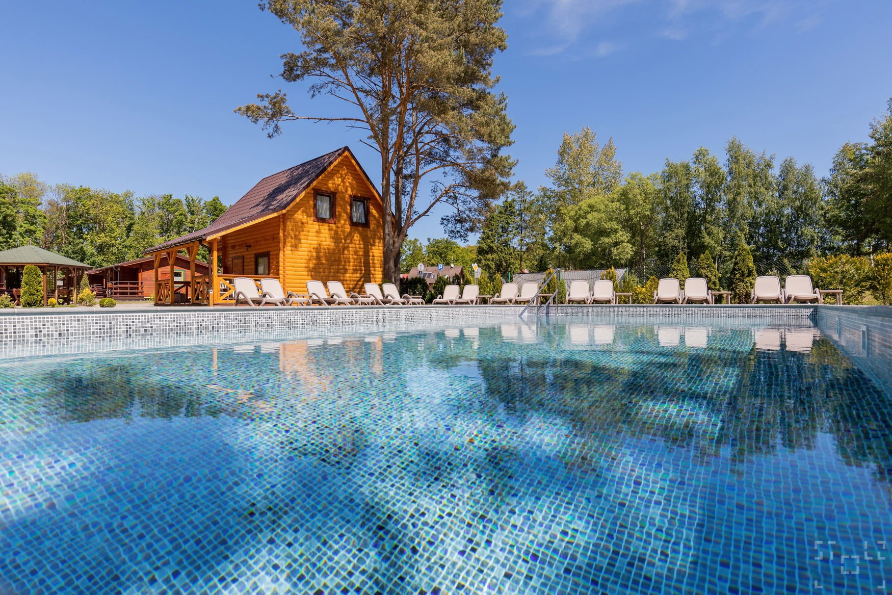 Błękitny basen z mozaikowym dnem, otoczony rzędem białych leżaków i drewnianymi budynkami w tle, sfotografowany w słoneczny dzień.