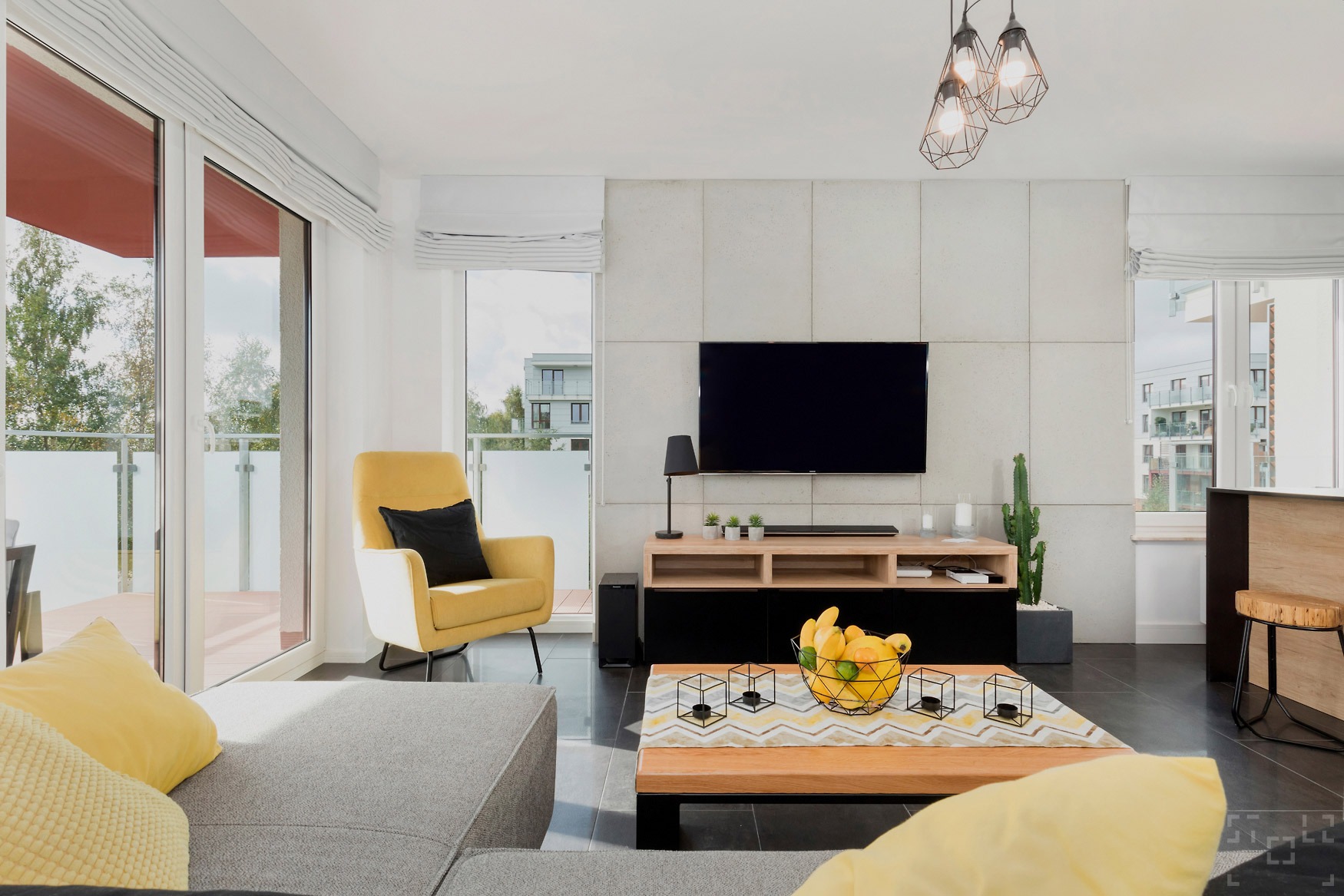 Jasne, nowoczesne wnętrze salonu z żółtym fotelem, szarą sofą, drewnianym stolikiem kawowym z owocami i telewizorem na ścianie z betonowych paneli.