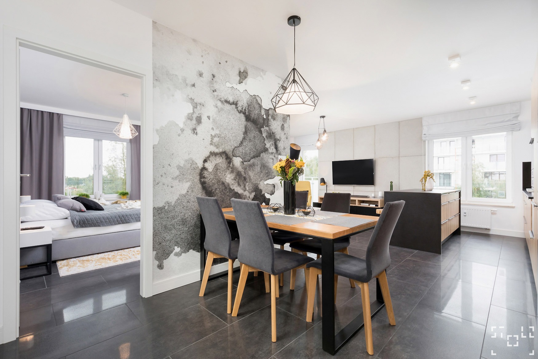 Jasne wnętrze mieszkania z otwartą przestrzenią: jadalnia z drewnianym stołem i szarymi krzesłami, ściana z wzorem akwareli, widok na sypialnię z szarym łóżkiem i zasłonami.